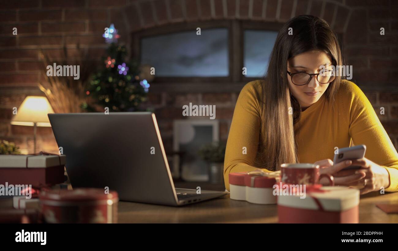 Junge Frau sitzt am Schreibtisch und plaudert mit ihrem Telefon an Heiligabend, an Feiertagen und am Technologiekonzept Stockfoto