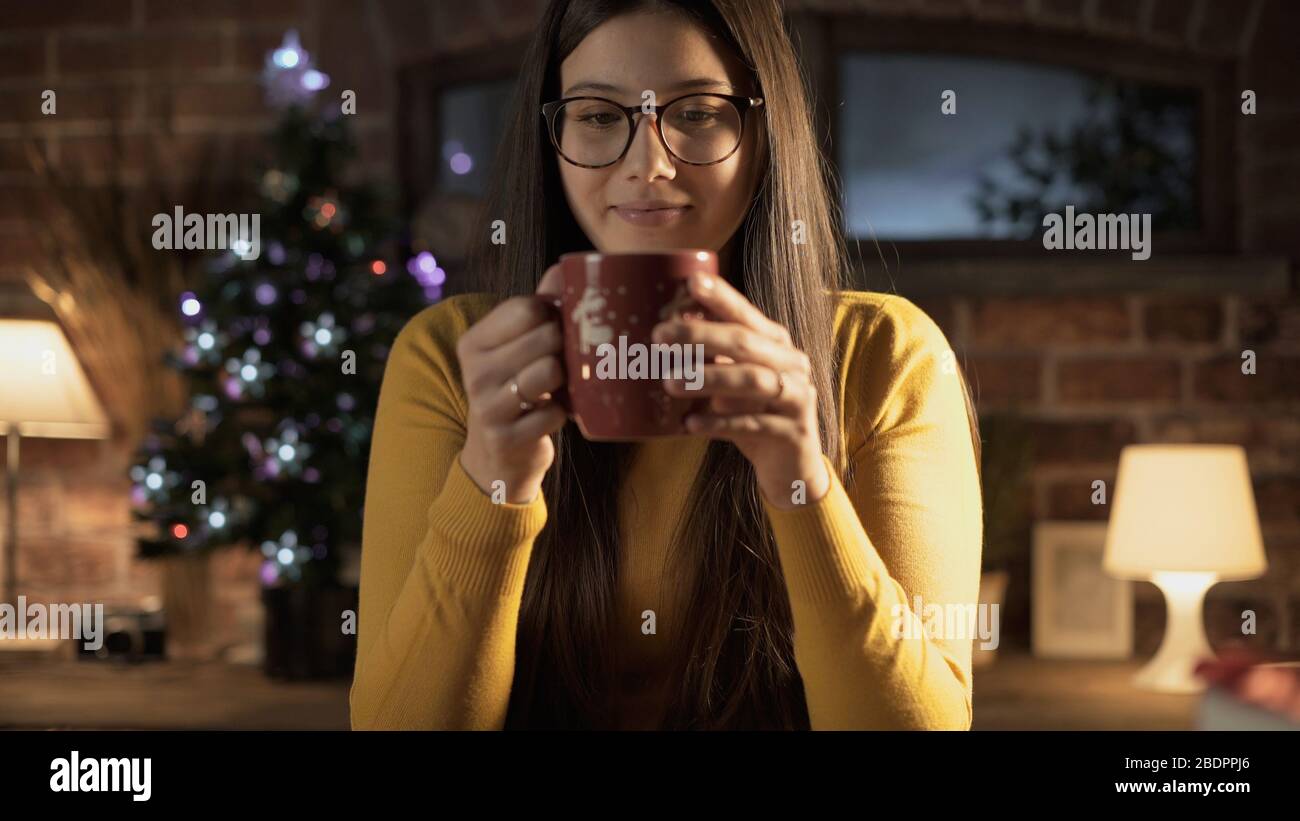 Fröhliche junge Frau, die zu Hause eine Tasse Tee hat, Weihnachtsbaum im Hintergrund, Weihnachtskonzept Stockfoto