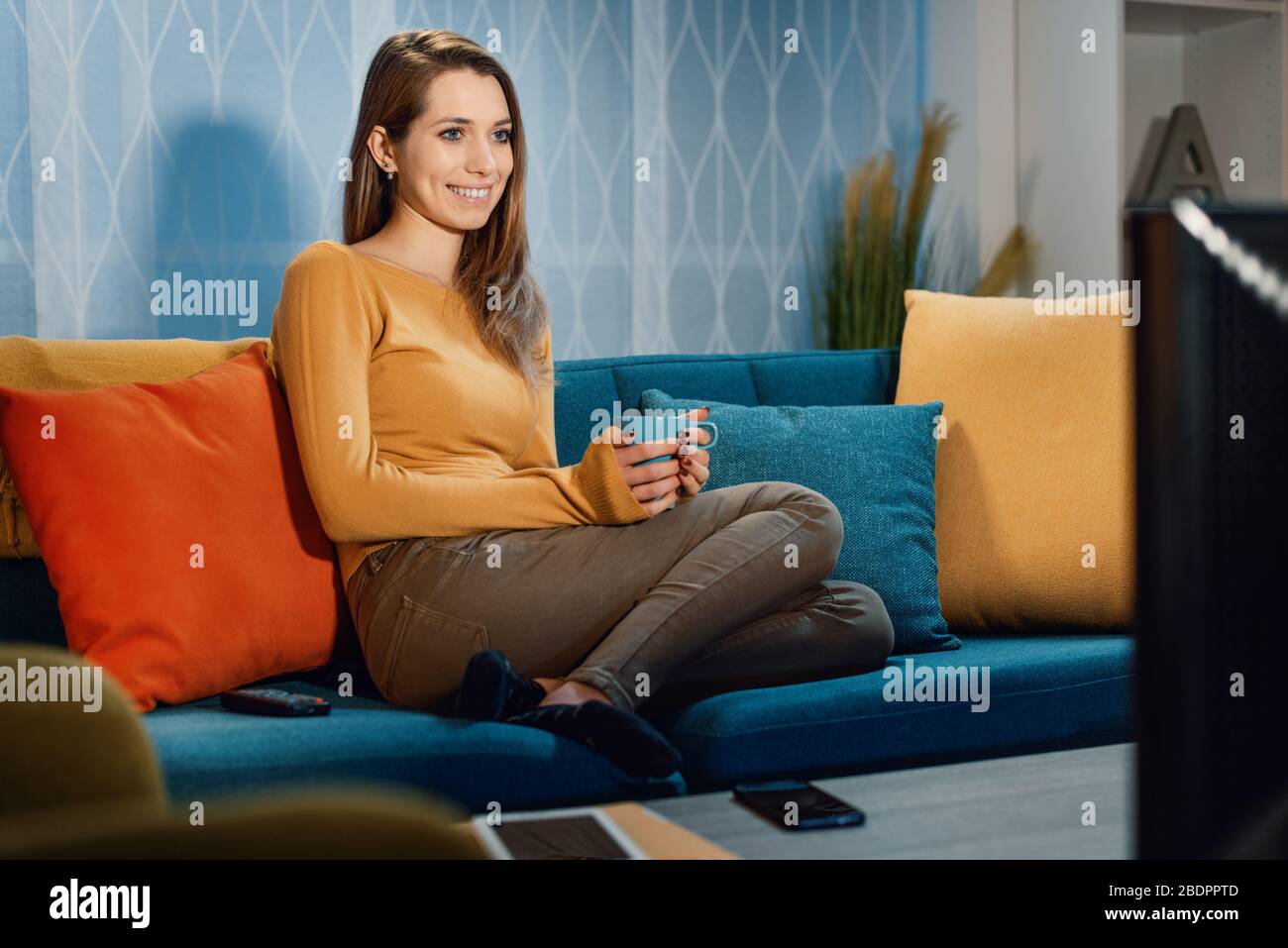 Fröhliche Frau, die ihren Lieblings-Telefilm im Fernsehen sieht, sitzt auf dem Sofa und hält eine Tasse Kaffee Stockfoto