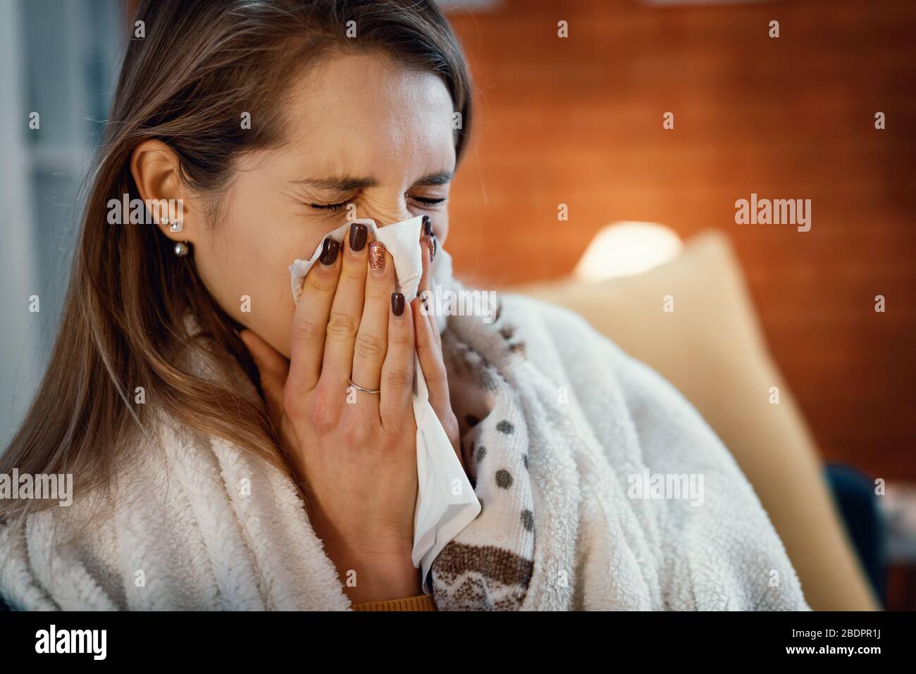 Junge Frau, die sich mit einer Decke bedeckt und ihre Nase weht, hat Kälte und Grippe Stockfoto