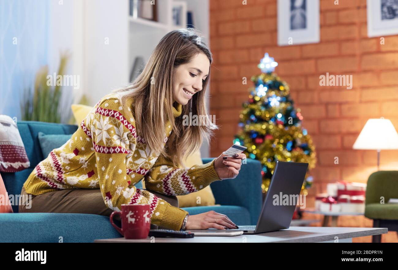 Fröhliche junge Frau sitzt zu Hause auf der Couch und macht Online-Shopping für Weihnachtsgeschenke, sie verbindet sich mit ihrem Laptop und hält einen Kredit c Stockfoto