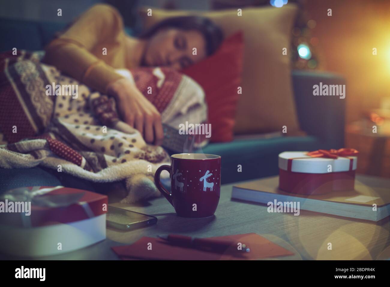 Junge Frau, die auf dem Sofa liegt und an Heiligabend einschläft, hält die Fernbedienung des Fernsehers, Geschenke im Vordergrund Stockfoto