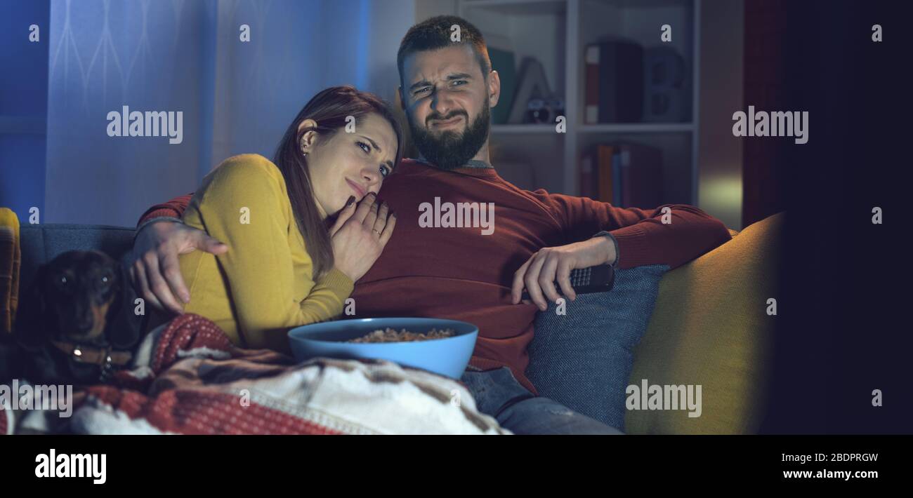 Das junge Paar sitzt zu Hause auf der Couch und sieht zusammen Horrorfilme im Fernsehen, er umarmt seine Freundin Stockfoto