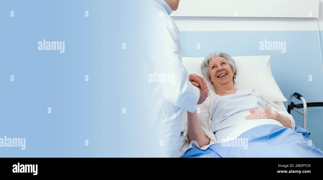 Doktor, der eine hospitalisierte Senioren-Patientin im Krankenhaus besucht, hält sich die Hand, lächelt und beruhigt ihr, Oberpflegekonzept Stockfoto