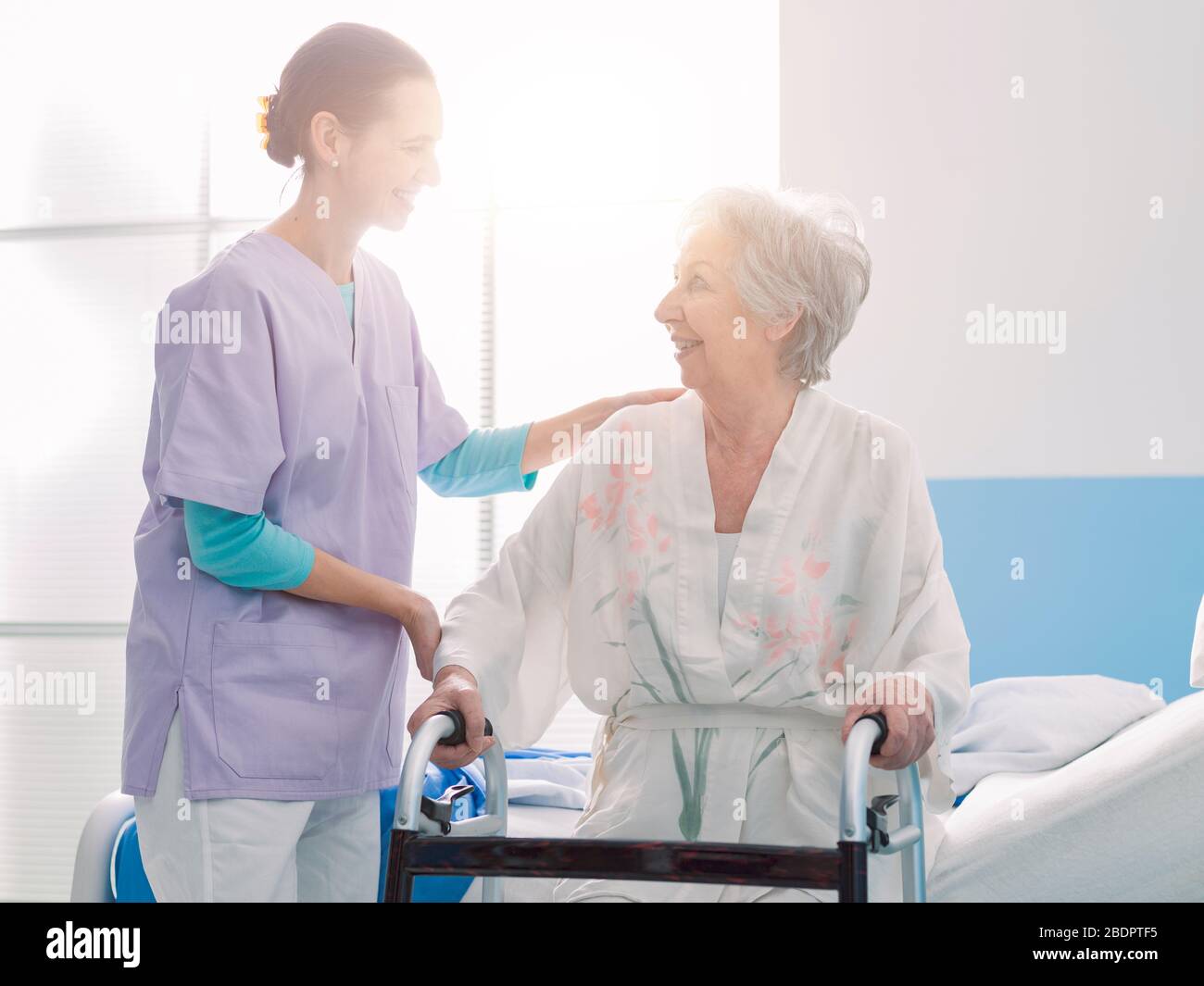 Lächelnde, professionelle Krankenschwester, die einen leitenden Patienten im Krankenhaus unterstützt, lehnt sich die Frau an einen Wanderer Stockfoto