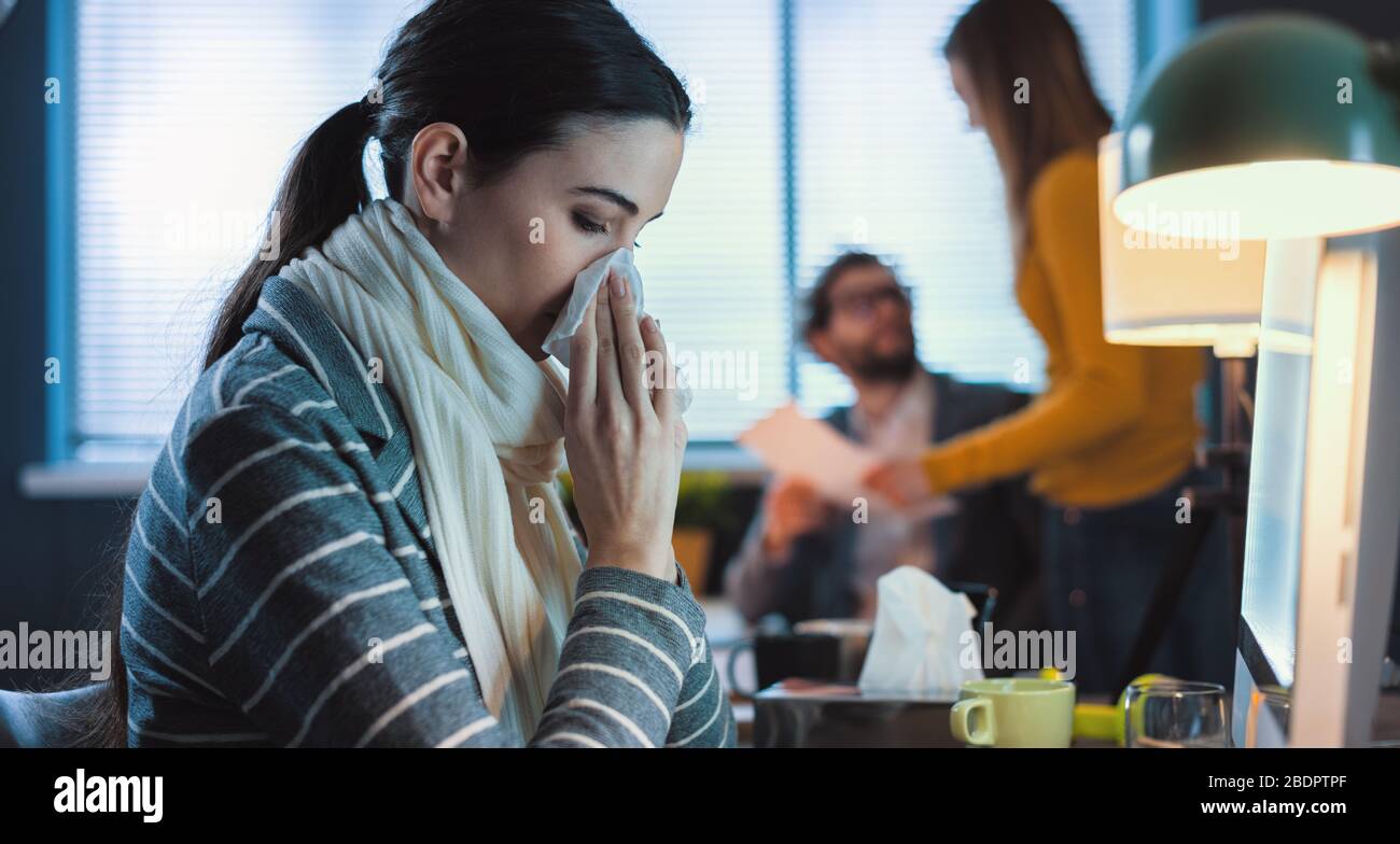 Junge Büroangestellte, die am Schreibtisch sitzen und arbeiten, hat sie eine Erkältung und bläst ihre Nase mit einem Gewebe Stockfoto