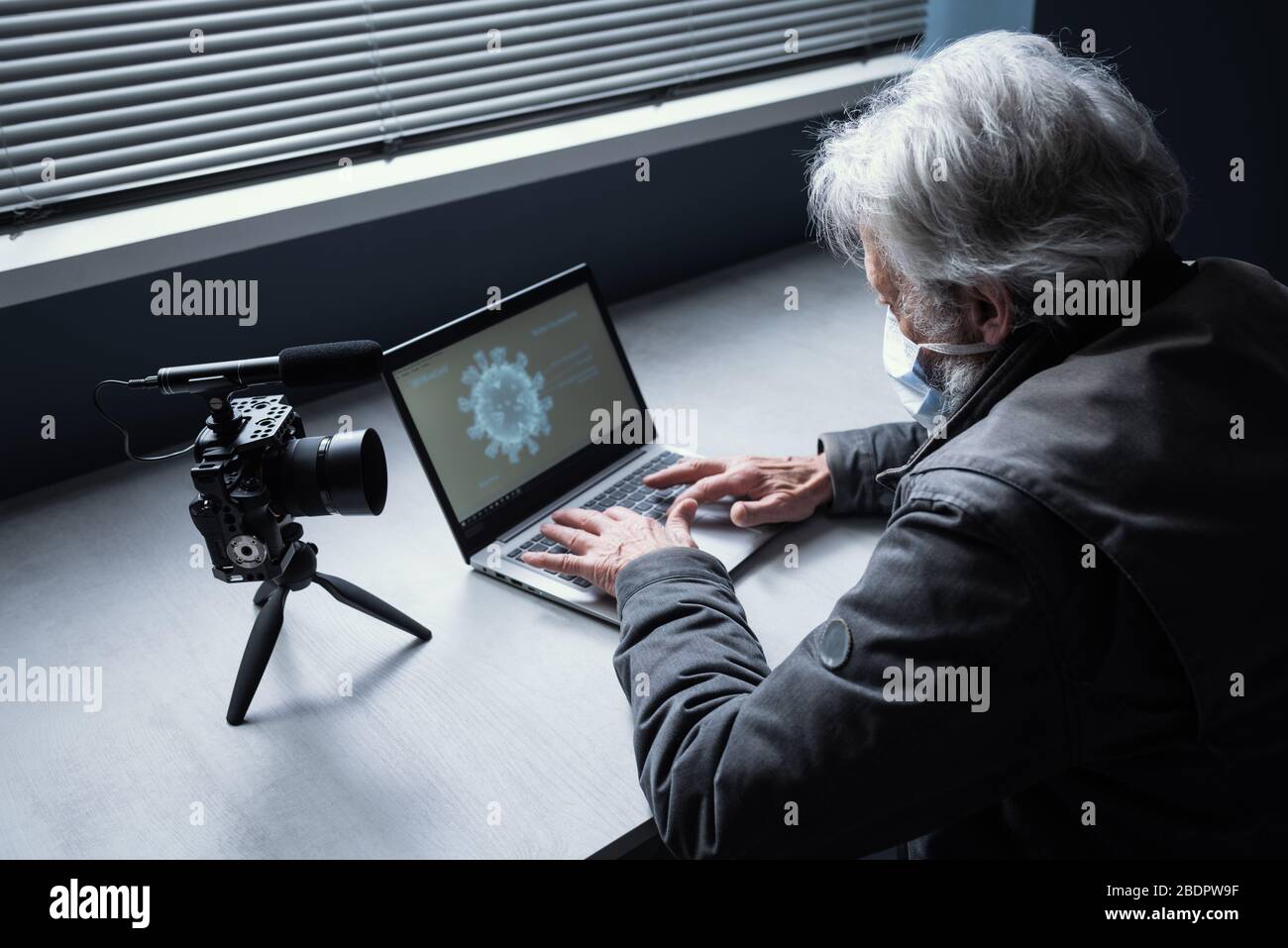 Senior man sitzt am Schreibtisch und trägt eine chirurgische Maske, er verbindet sich mit seinem Laptop und streamt ein Video online auf seinem Kanal, Covid 19 Virus o Stockfoto