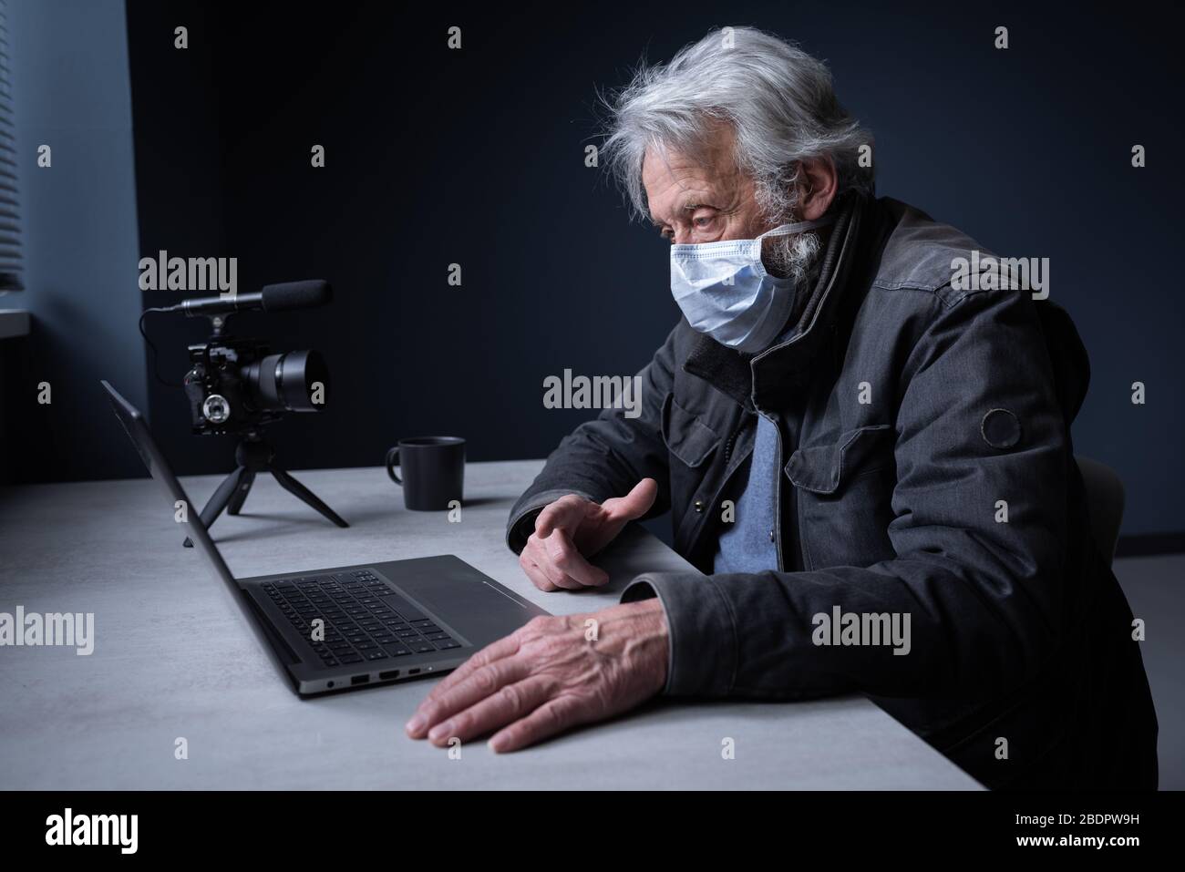 Senior man sitzt am Schreibtisch und trägt eine chirurgische Maske, er verbindet sich mit seinem Laptop und streamt ein Video online auf seinem Kanal, Covid 19 Virus o Stockfoto