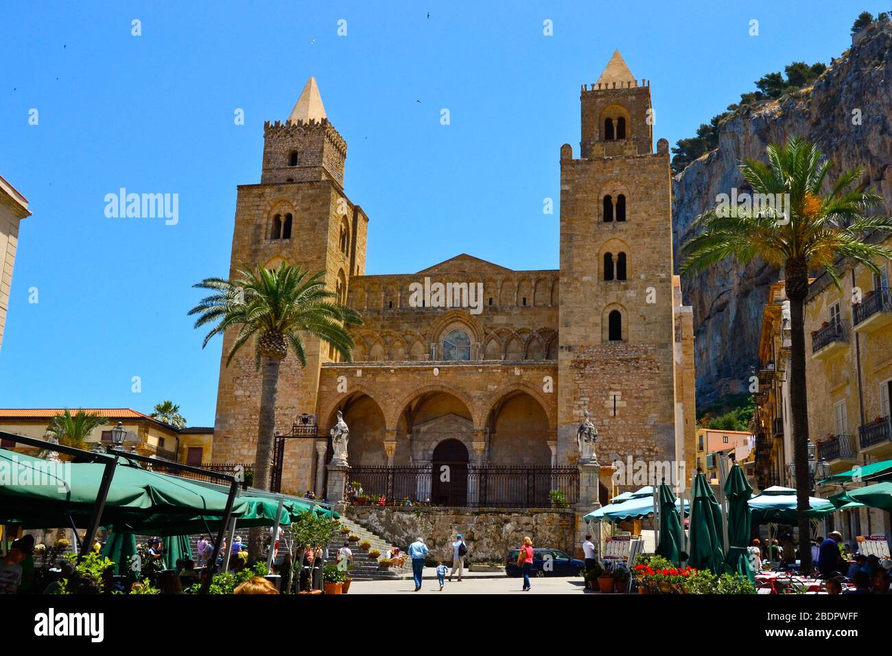 Cafalu Kathedrale aus goldenen Ziegeln, zwei Türmen und normannischen Stil mit Palmen und grünen Sonnenschirmen unter einem strahlend blauen Himmel an einem sonnigen Tag Stockfoto