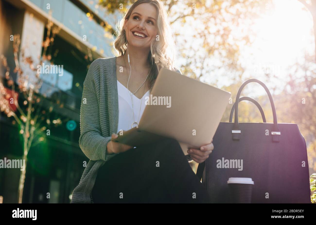 Frau mit Ohrhörern, die draußen mit Laptop sitzt. Geschäftsfrau sitzt draußen in der Stadt mit Laptop an einem sonnigen Tag. Stockfoto