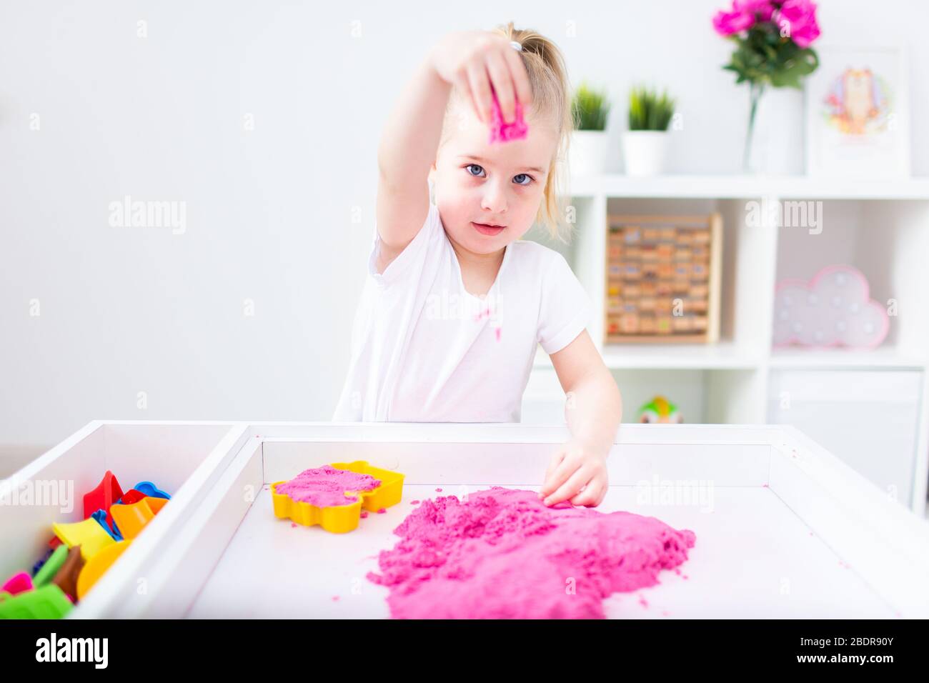 Kleines blondes Mädchen spielt mit rosa kinetischen Sand auf einem weißen Tisch in einem hellen Raum. Sensorische Entwicklung. Unterricht in einem Kindergarten Stockfoto