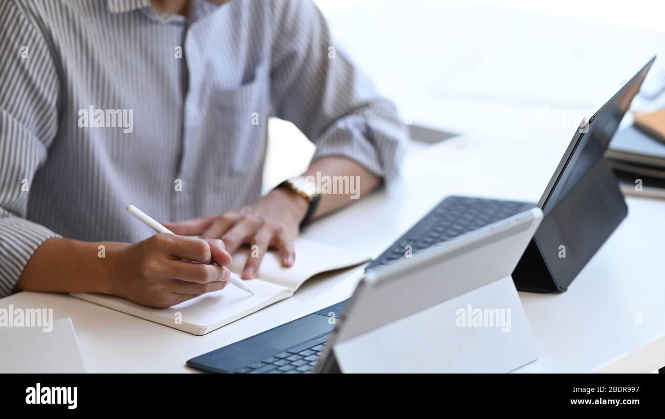 Bild von smart Mann arbeitet als Project Administrator Schreiben auf Notebook während sitzen vor Computer-Tablet mit Tastatur-Hülle mit dem Mod abgeschnitten Stockfoto