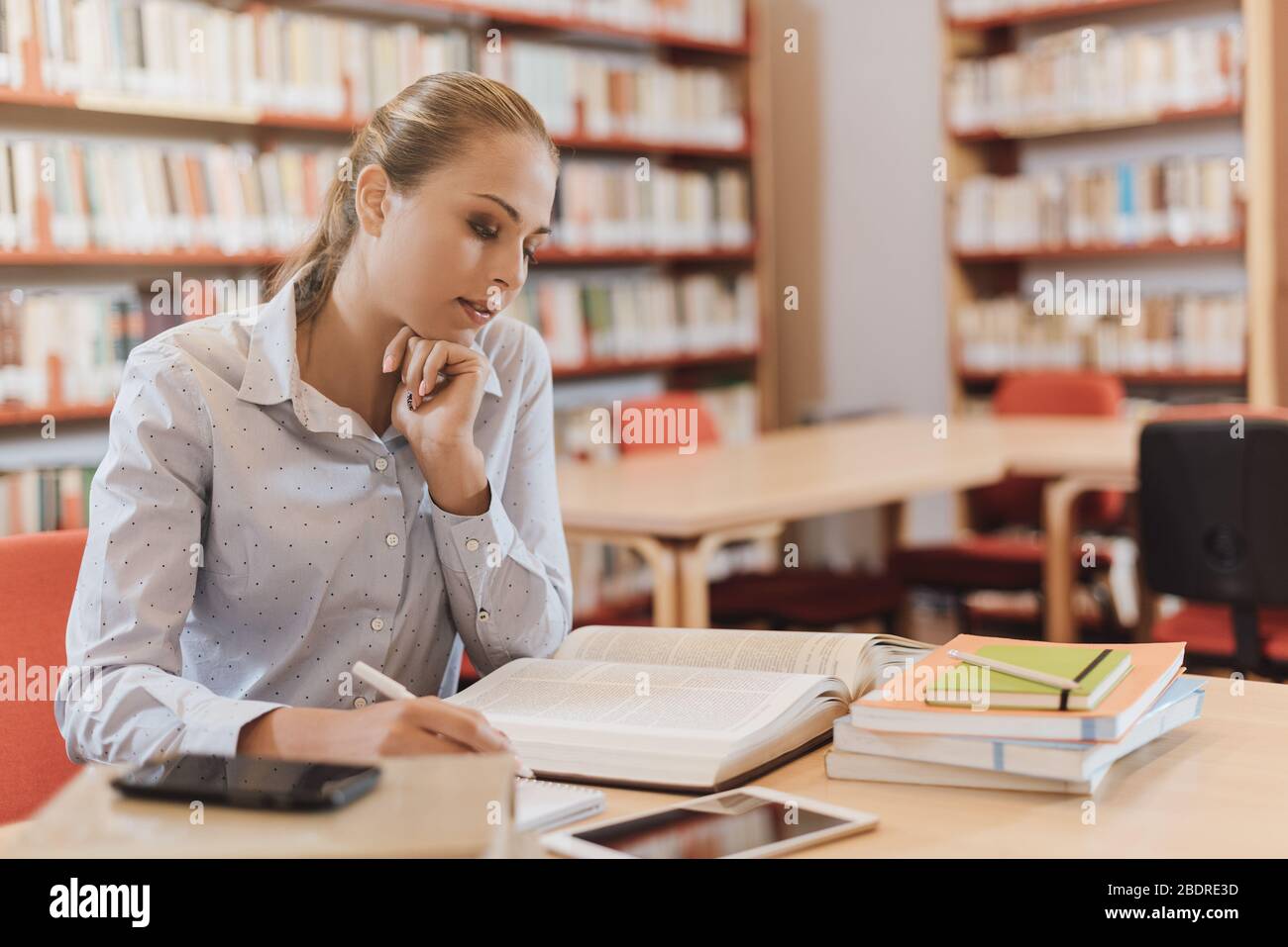 Effiziente Studentin in der Bibliothek sitzt sie am Schreibtisch und konzentriert sich auf Studium und Lernen, Bücherregal im Hintergrund Stockfoto
