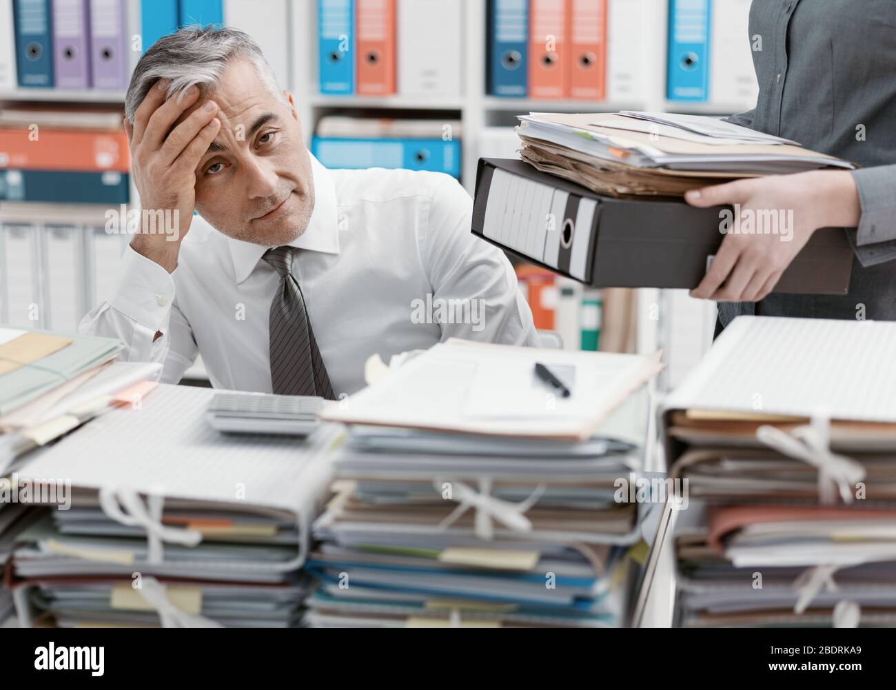 Betonte Geschäftsmann im Büro Schreibtisch arbeiten und mit Arbeit überlastet, der Desktop mit Papierkram abgedeckt ist, seine Sekretärin bringt mehr Dateien Stockfoto