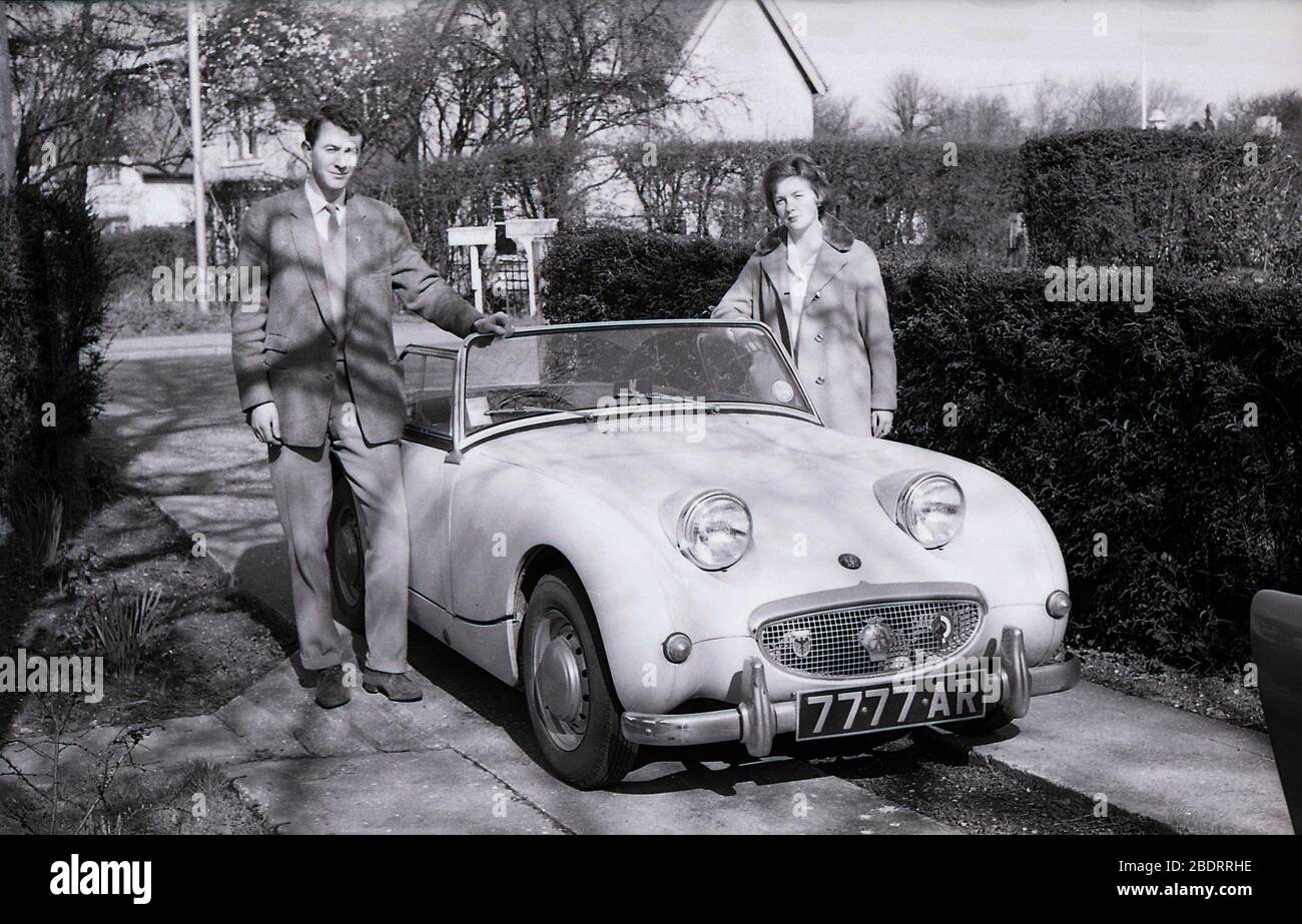 1960er Jahre, historisch, ein junges erwachsenes Paar, das neben ihrem offenen Sportwagen Austin-Healey Sprite stand, parkte neben einer Hecke auf einer schmalen Betonzufahrt im Eastholm Green in Letchworth Garden City, England, Großbritannien. Der kleine Zweisitzer Sprite, der als Nachfolger des Vorkriegs Austin Seven entworfen wurde, wurde zwischen 1958-1961 in Großbritannien produziert und mit seinen Scheinwerfern, die aus der Fronthaube kommen, wurde er liebevoll als "Frogeye" bekannt. Stockfoto