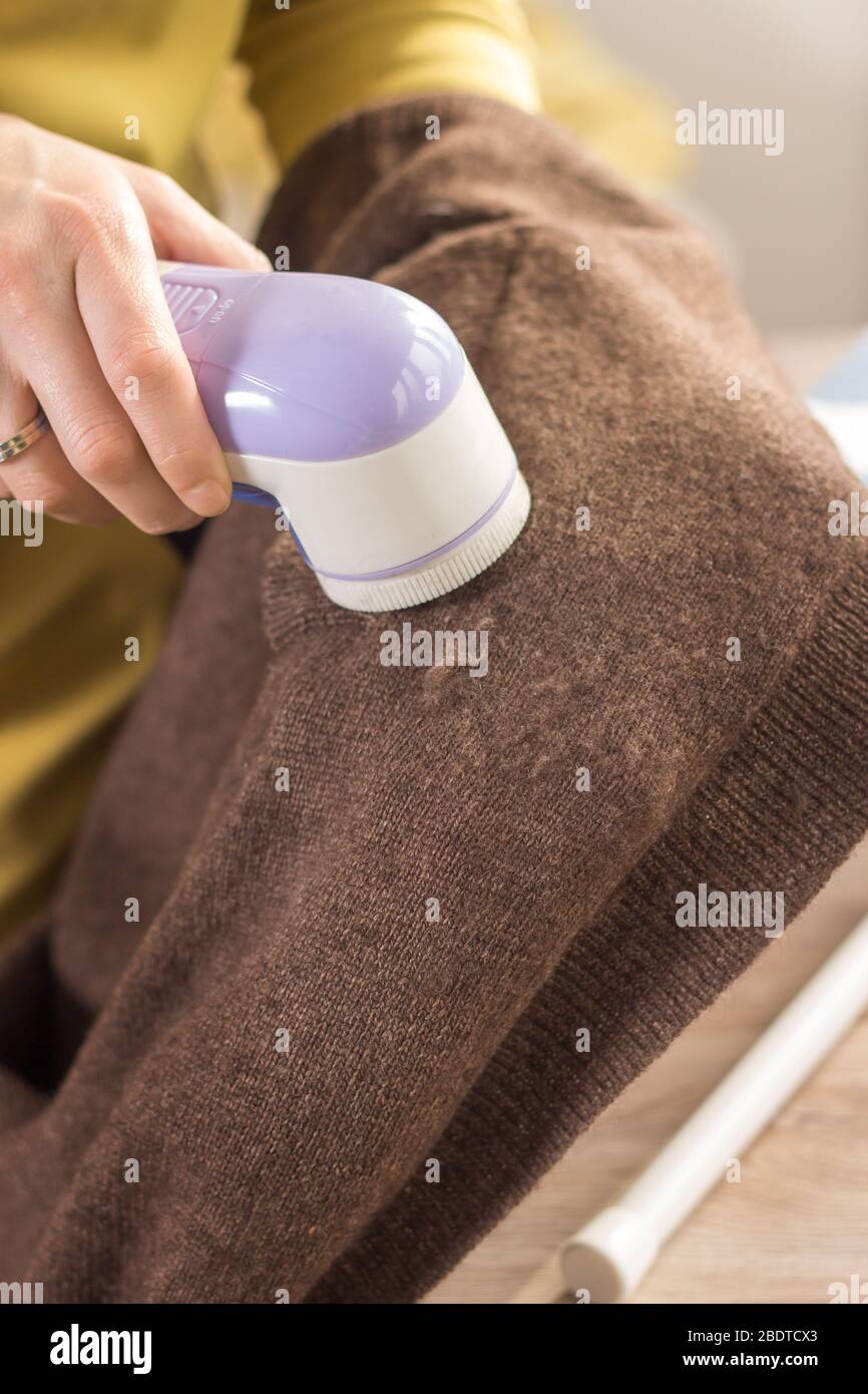 Die Frau hält einen Elektrorasierer in der Hand, um melierte Kleidung zu reinigen und reinigt den Ärmel eines braunen Wollpullovers. Stockfoto