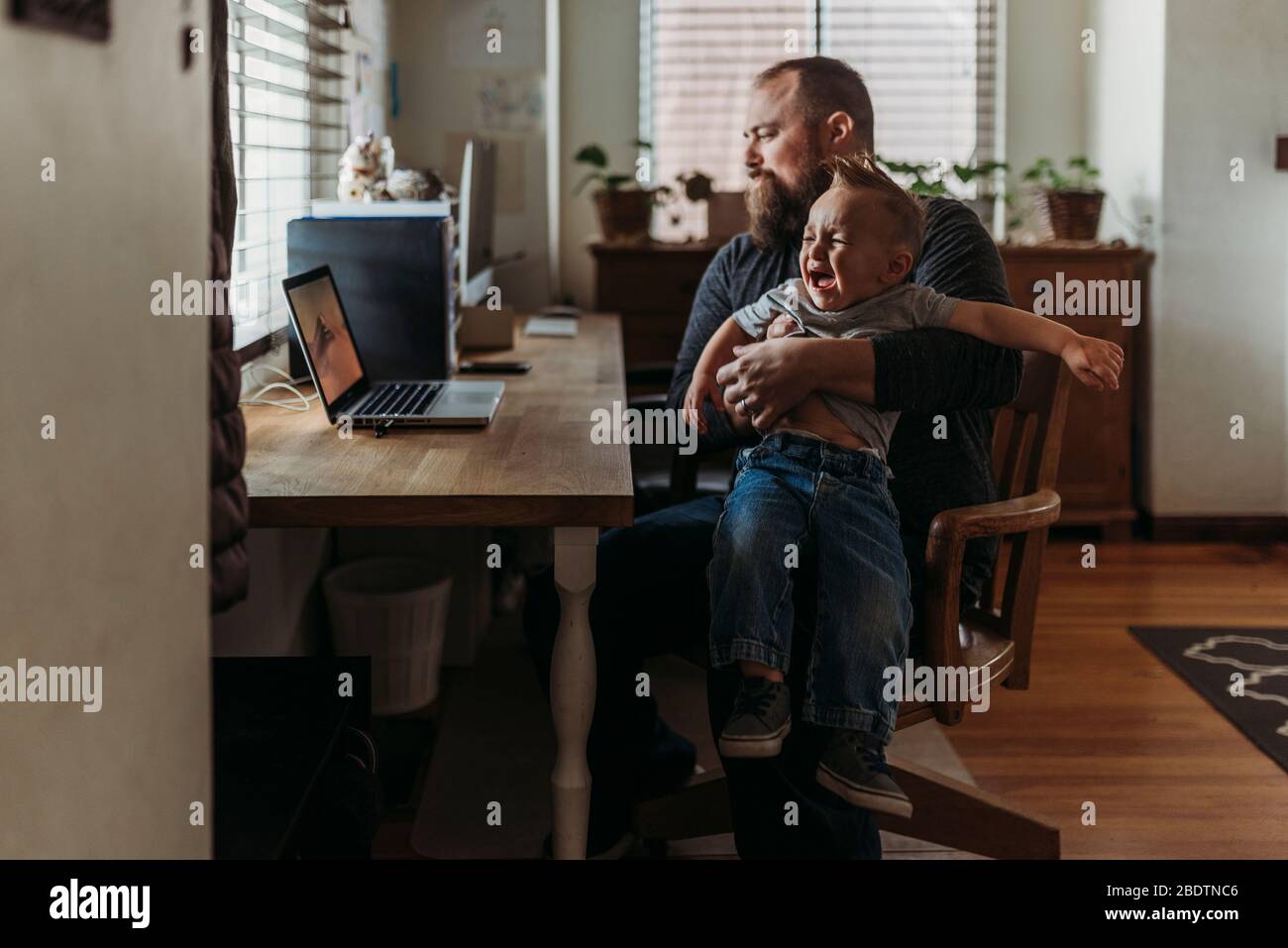 Vater arbeitet von zu Hause mit einem einjährigen Jungen weinend in seinem Schoß Stockfoto