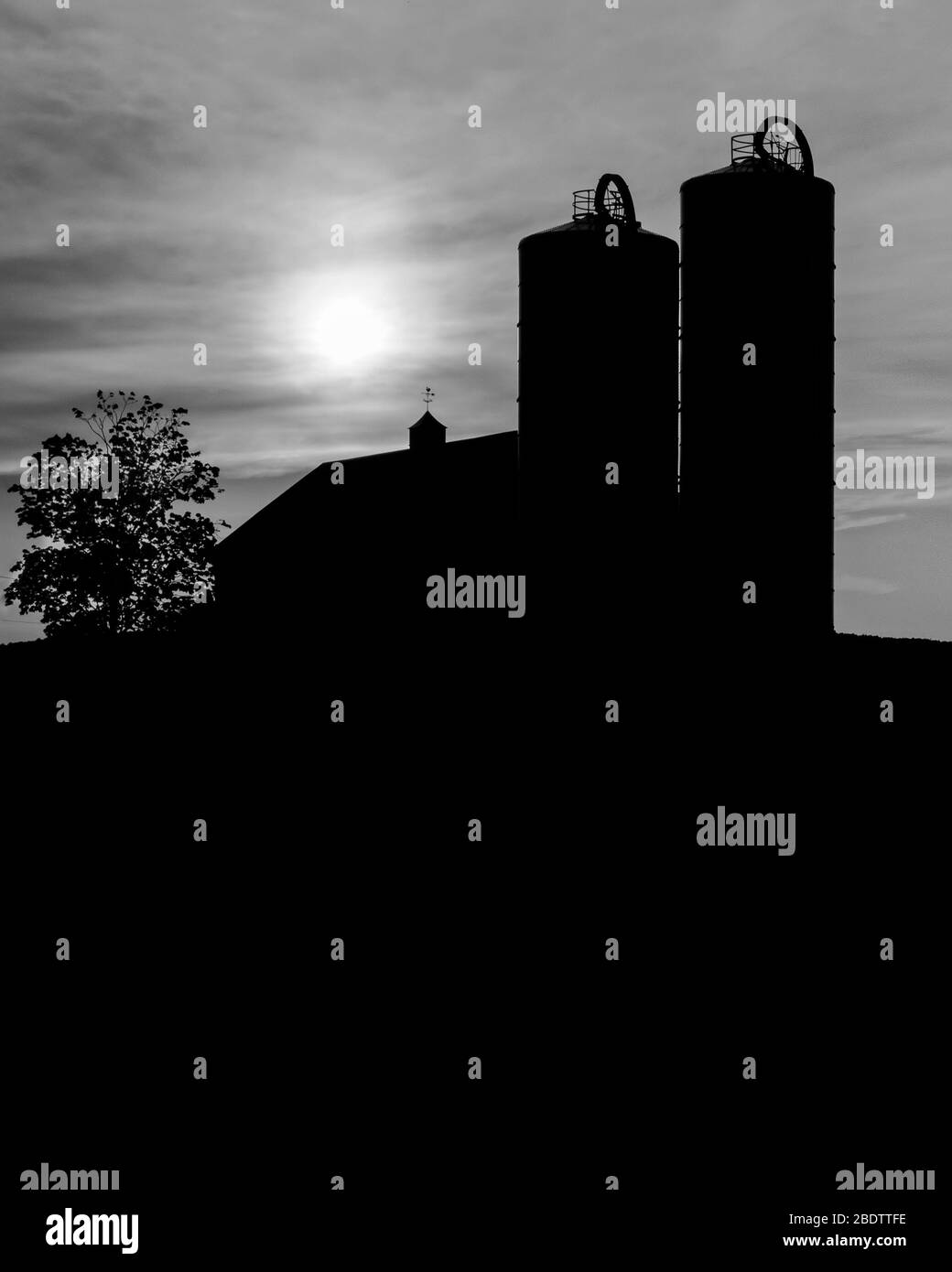 Zwei Silos auf einem Bauernhof in schwarz-weißer Silhouette mit einer verblassten Sonne hinter den Wolken. Stockfoto