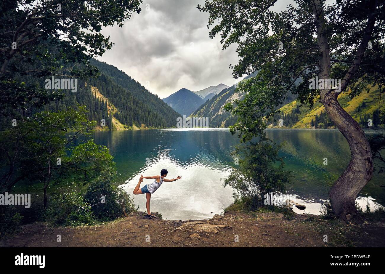 Junge Frau ist Yoga balance Pose am Bergsee mit bedecktem Himmel Hintergrund Stockfoto