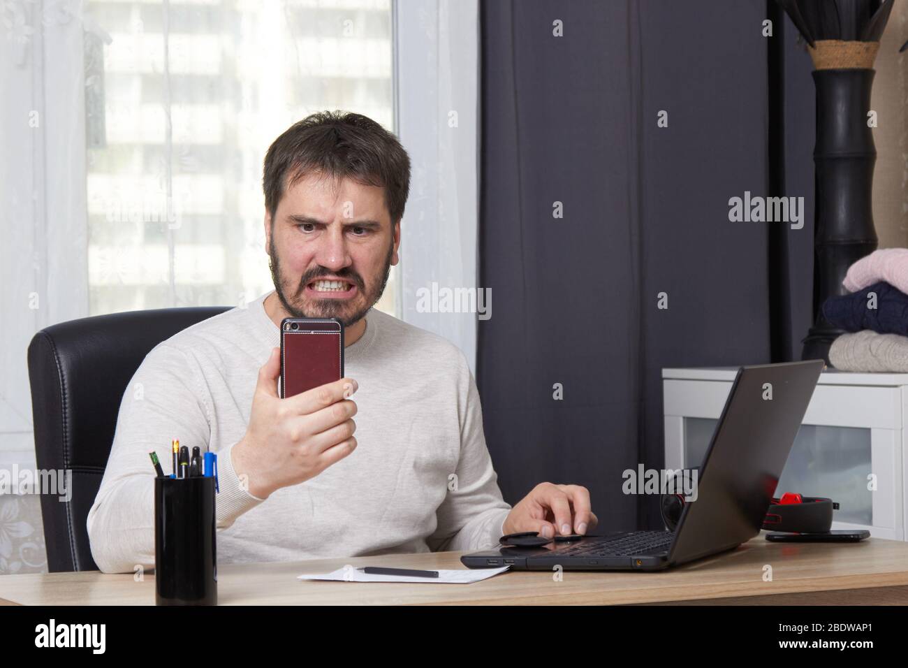 Ein bärtiger Mann sitzt an einem Tisch mit einem Laptop in einem weißen Pullover, der nach einem Job sucht, der auf einem Handy anruft Stockfoto