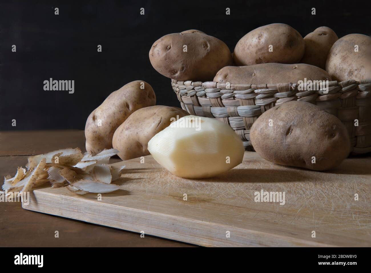 Geschälte Kartoffeln auf einem Schneidebrett mit mehr Kartoffeln in einem Korb im Hintergrund in der niedrigen Licht. Stockfoto