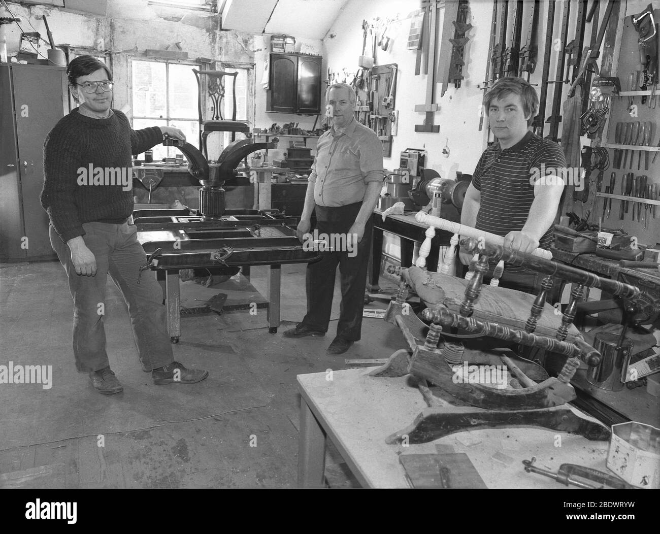 1980er Jahre, historisch, drei Möbelrestauratoren oder Restauratoren, die in ihrer Werkstatt stehen, die Wände sind mit Werkzeugen gesäumt und mit einem antiken Tisch und einem kleinen Hocker, der auf Reparatur und Restaurierung wartet, England, Großbritannien. Die Restaurierung alter und antiker Möbel ist ein Fachmann und Fachmann, da jedes Stück unterschiedliche Restaurierungsbedürfnisse hat. Stockfoto