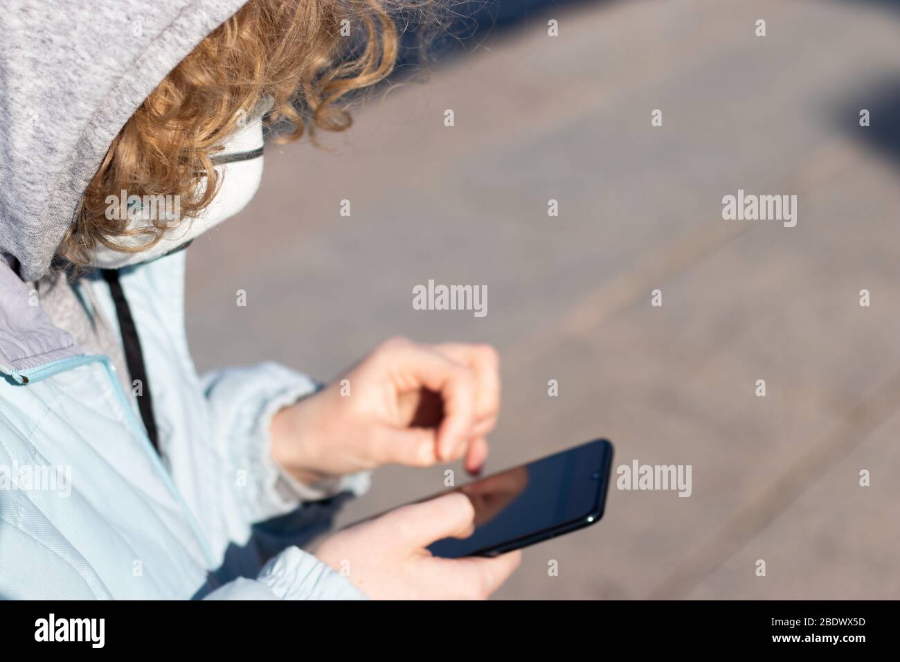Frau in Kapuze und Jacke benutzt ein Telefon auf der Straße. Frau mit medizinischer Schutzmaske. Das Konzept des technologischen Fortschritts in Stockfoto