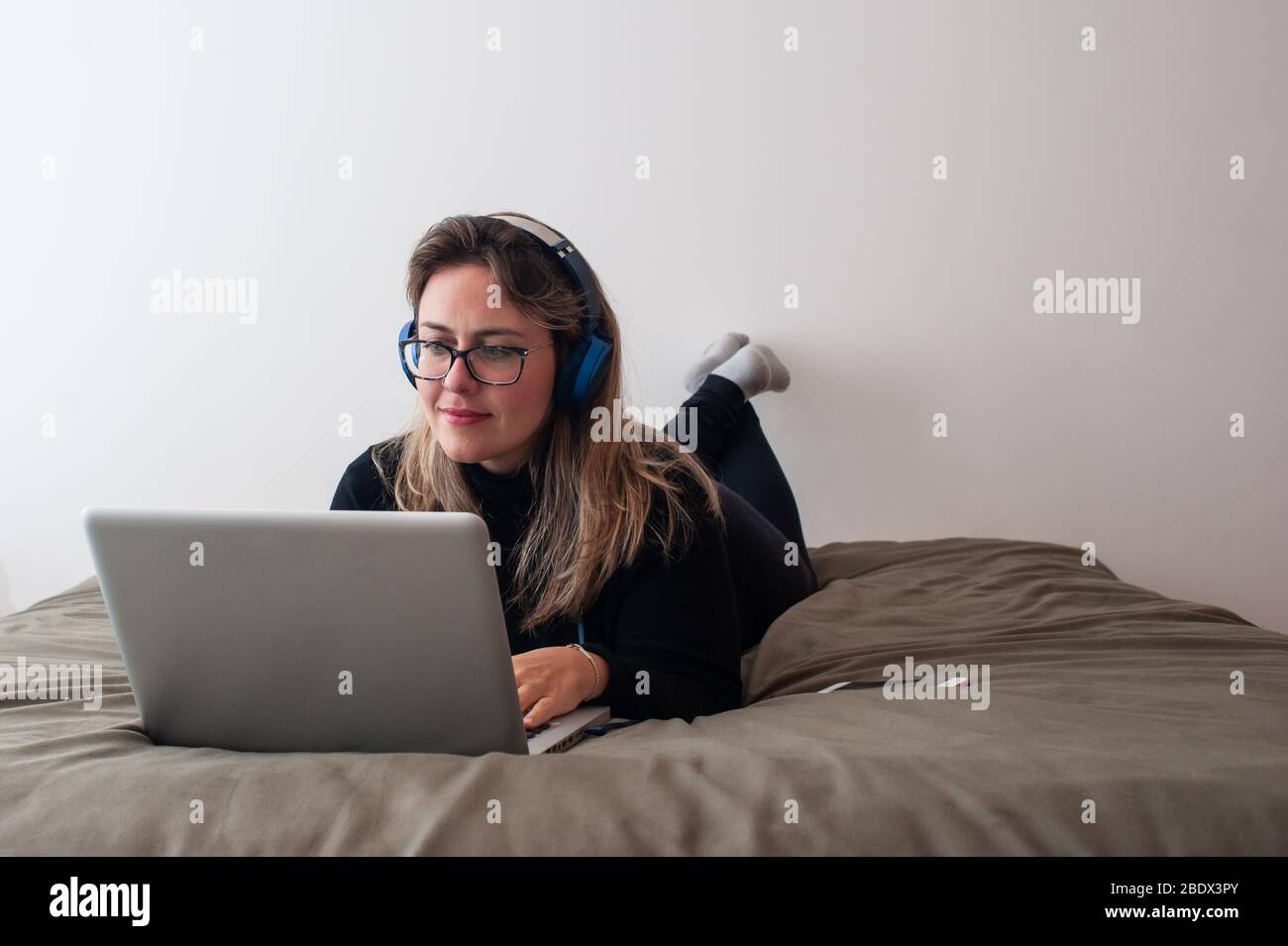 Blonde Frau, die sich im Bett mit dem Computer hinlegt. Sie trägt Brillen und benutzt einen Kopfhörer Stockfoto
