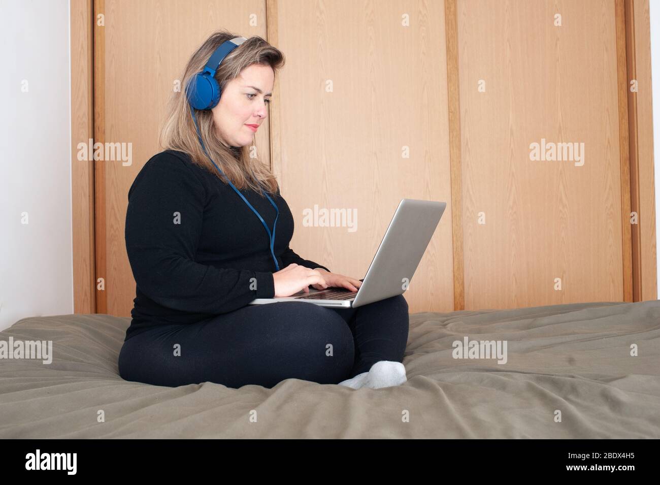 Blonde Frau sitzt im Bett mit dem Computer. Sie ist mit einem Kopfhörer Stockfoto