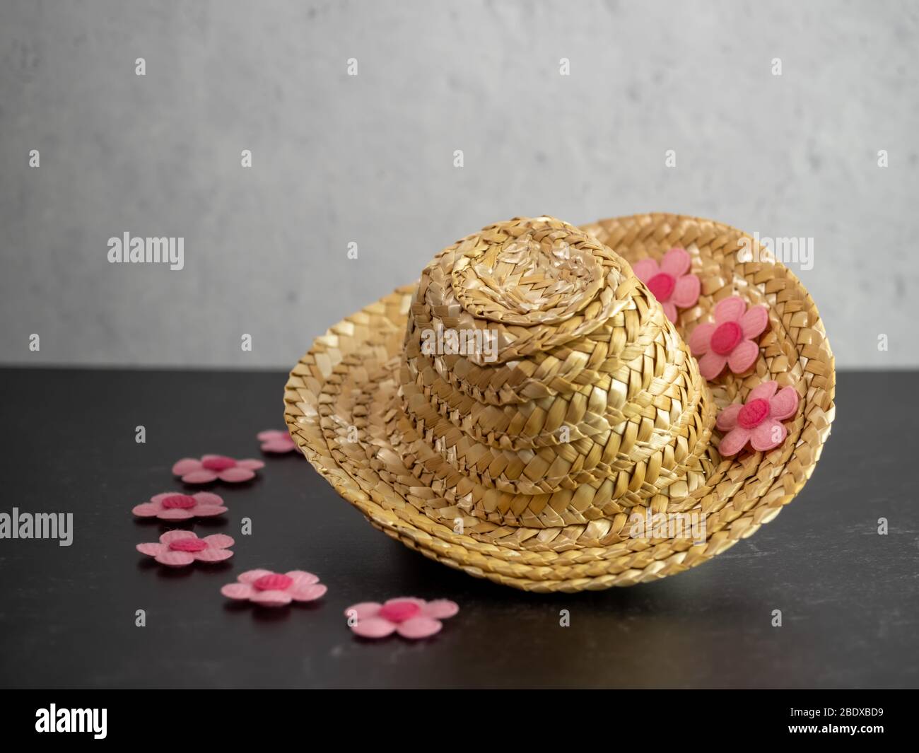 Stroh Kuh Mädchen Hut sitzt auf einem schwarzen Schiefer Zähler mit rosa Filzblumen schmücken es und eine graue Gipswand. Mode-Accessoire mit Blumen. Stockfoto
