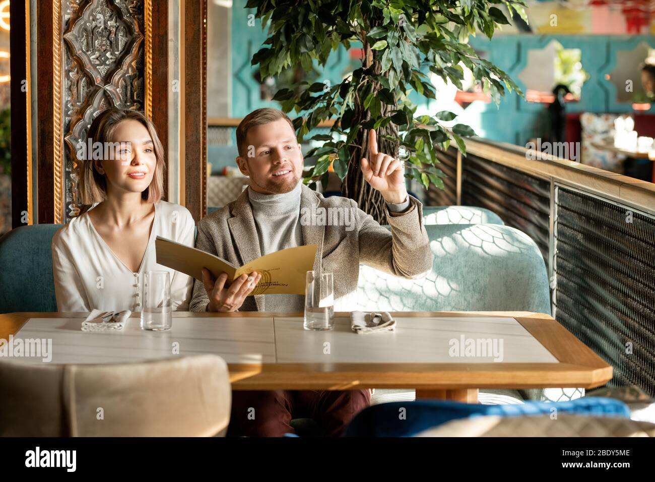 Junger Mann mit erhöhtem Zeigefinger, der darum bittet, dass der Kellner im Restaurant Ordnung bringen kann, während er neben seiner Freundin auf der Couch sitzt Stockfoto