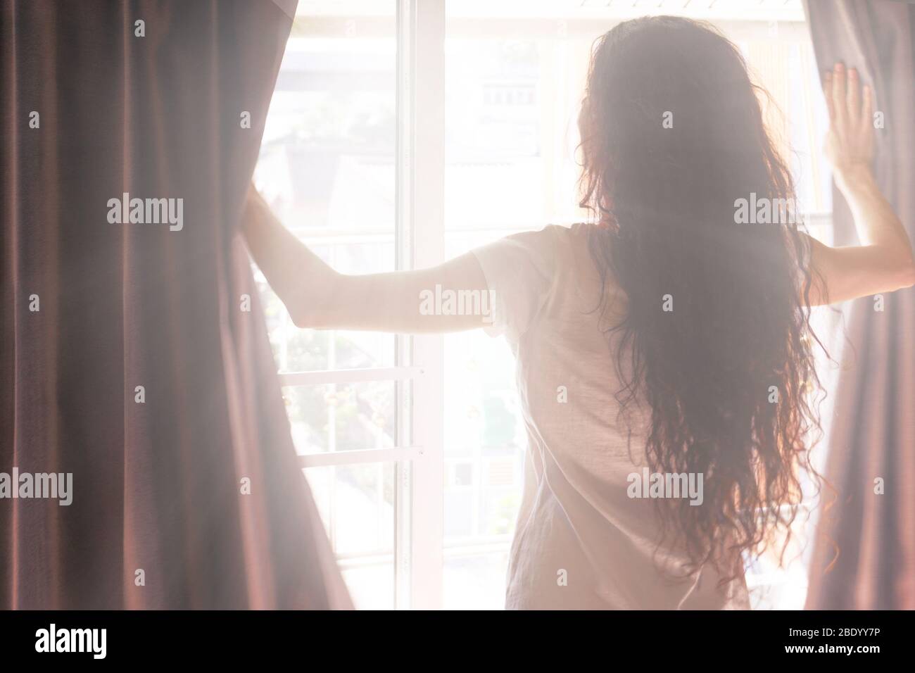 Sonniger Morgen - eine Frau öffnet die Vorhänge und die Sonne scheint durch das Fenster. Rückansicht. Verschwommenes Foto Stockfoto
