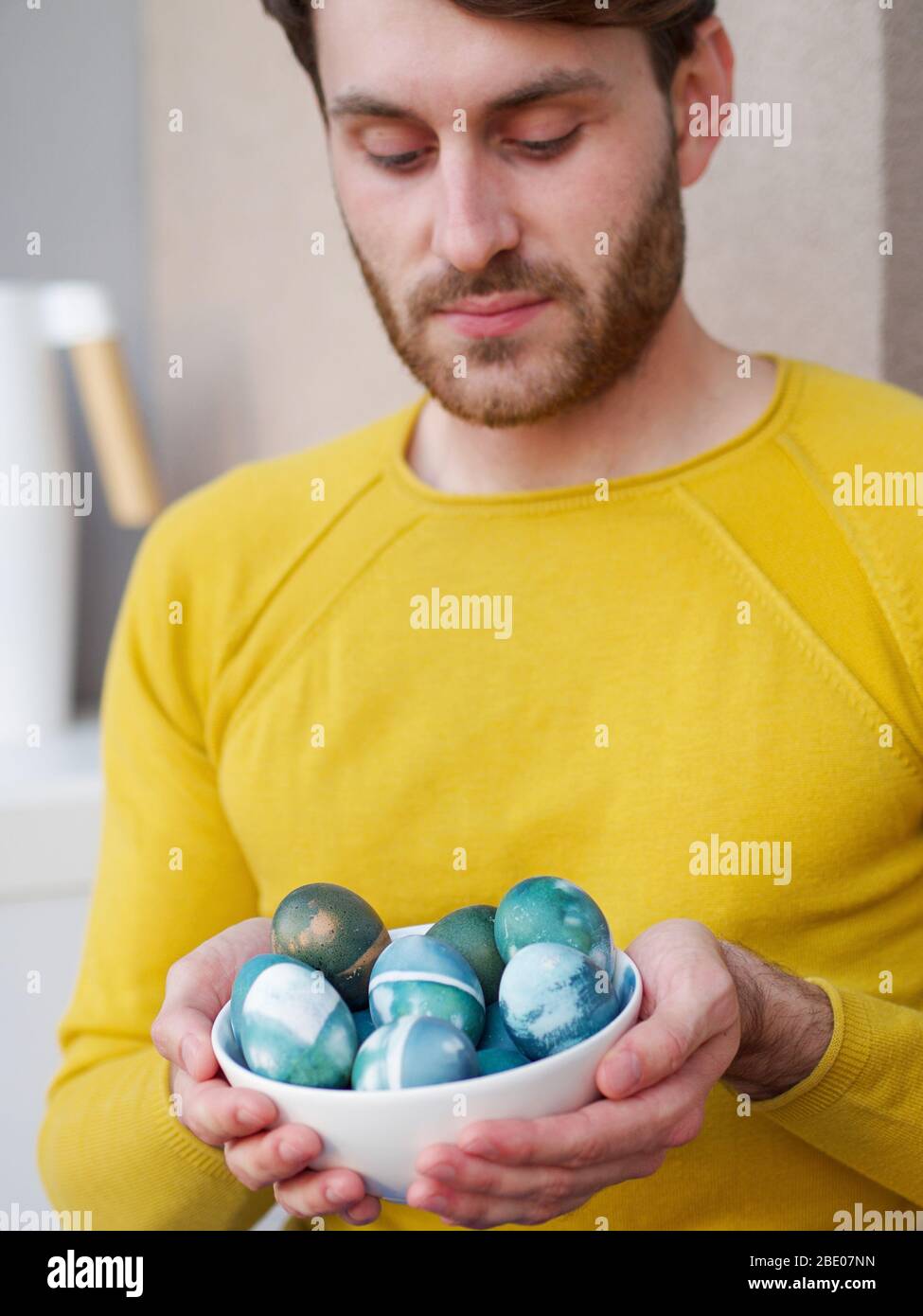 Kaukasischer Mann hält eine blaue ostereier in einer Schüssel während der osterzeit, mit natürlichen Rotkohl Färbung gefärbt, trägt eine gelbe sweather. Stockfoto