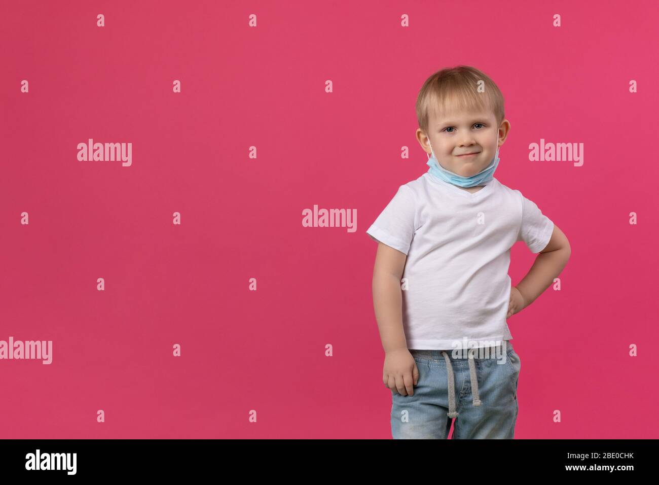 Ein blondes Knabenkind steht auf einem leuchtend rosa Hintergrund, Hand an Seite und sieht spöttisch und sarkastisch auf die Kamera, eine Maske wird auf sein Gesicht gelegt. Clo Stockfoto