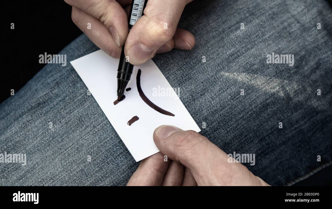 Schreiben sehr traurig Lächeln Emoji auf weißem Papier Karte in Händen auf Jeans Textur Hintergrund. Traurige Stimmung schlecht farblose Zeit Stockfoto