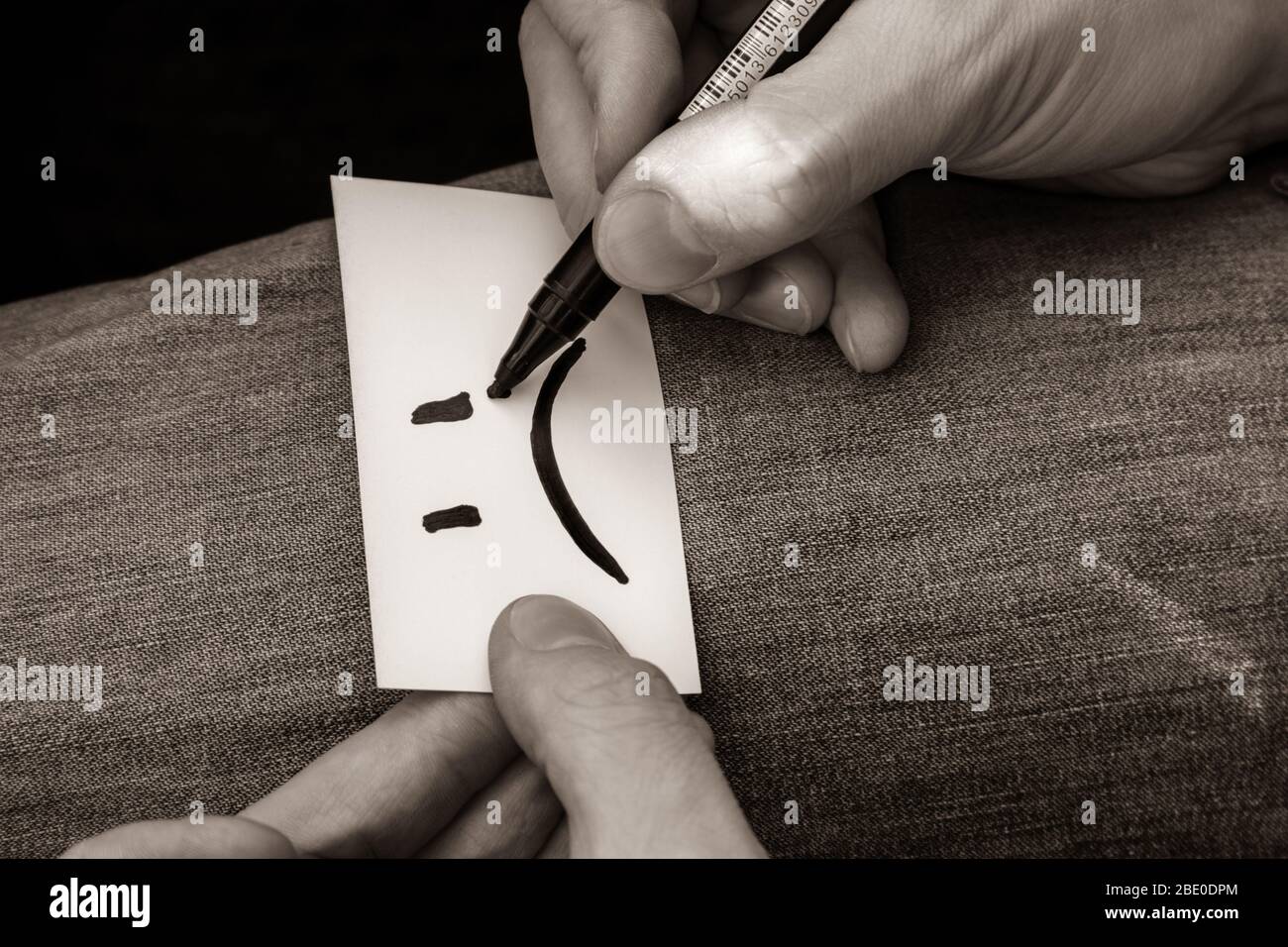 Schreiben sehr traurig Lächeln Emoji auf weißem Papier Karte in Händen auf Jeans Textur Hintergrund. Traurige Stimmung schlecht farblose Sepia Zeit Stockfoto