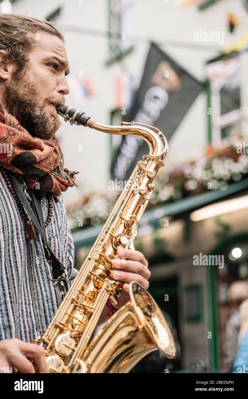 Vertikales Foto eines Saxophonisten bärtig und mit blonden Haaren spielend mit hellen Emotionen auf der Straße voller Menschen Stockfoto