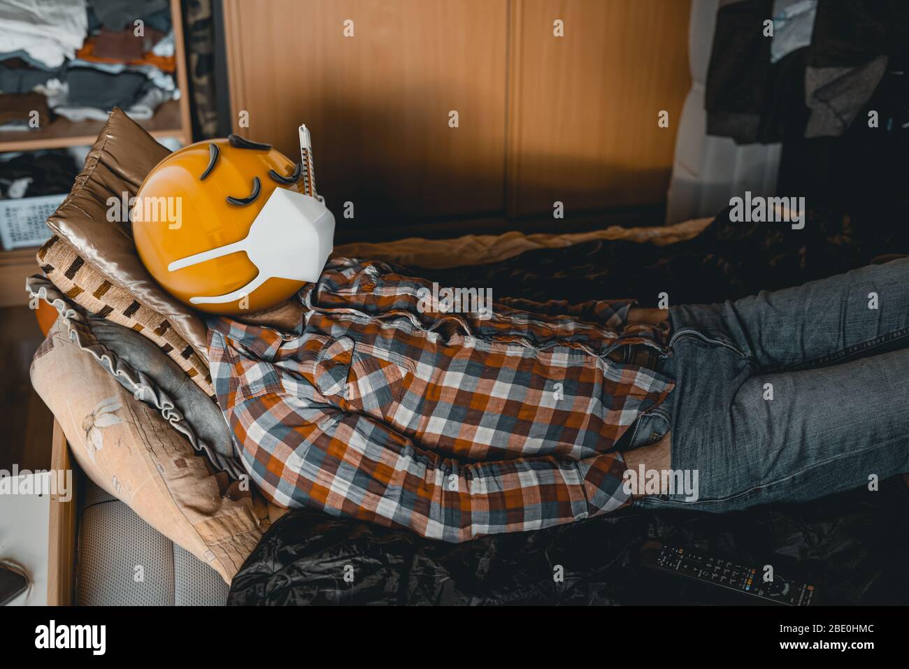 Kranker Mann mit Foto realistische gelbe Emoji Gesicht liegt auf einem Bett mit Thermometer in seinem Mund und Maske, bedeckt seine muth Schutz gegen Coronavirus Stockfoto