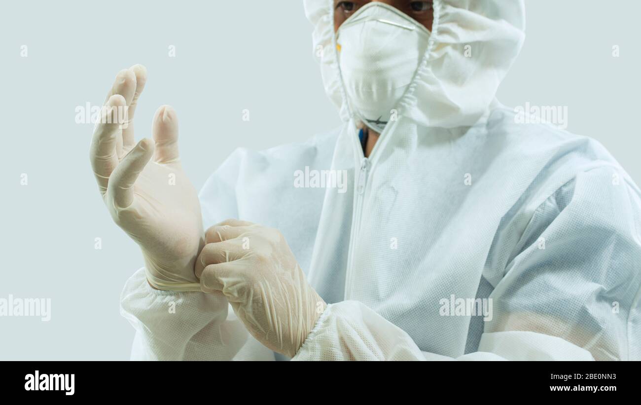 Arzt mit Maske und weißem Bioprotective Anzug, der Latexhandschuhe auf die Hände auf weißem Hintergrund legt Stockfoto