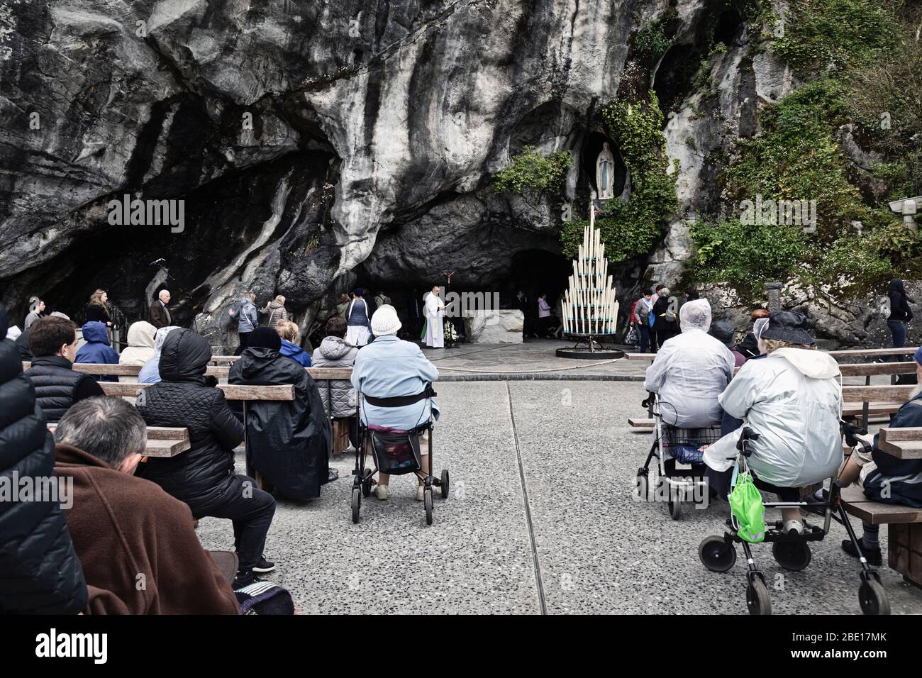 Ein Priester betet am Eingang der Massabielle Grotte des Heiligtums unserer Lieben Frau von Lourdes, während die Gläubigen Schlange stehen, um einzutreten. Das war es Stockfoto