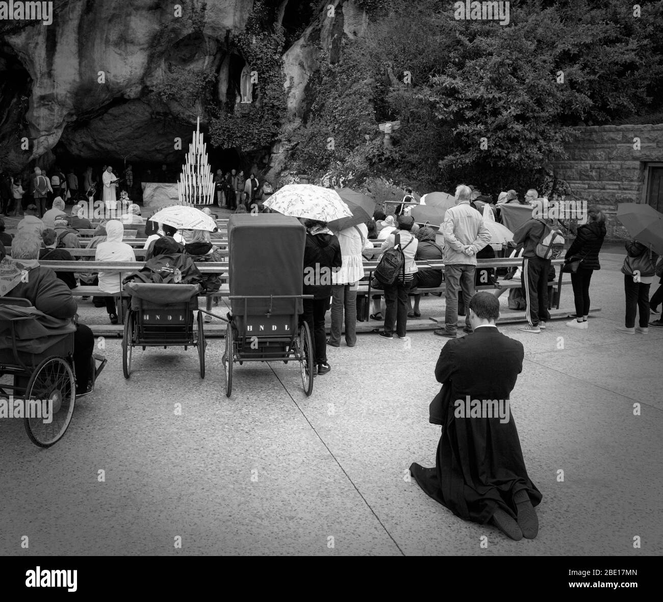 Ein Priester kniet zum Beten. Vor ihm Menschen im Rollstuhl. Im Hintergrund rezitiert ein anderer Priester Gebete am Eingang der Massabielle Stockfoto