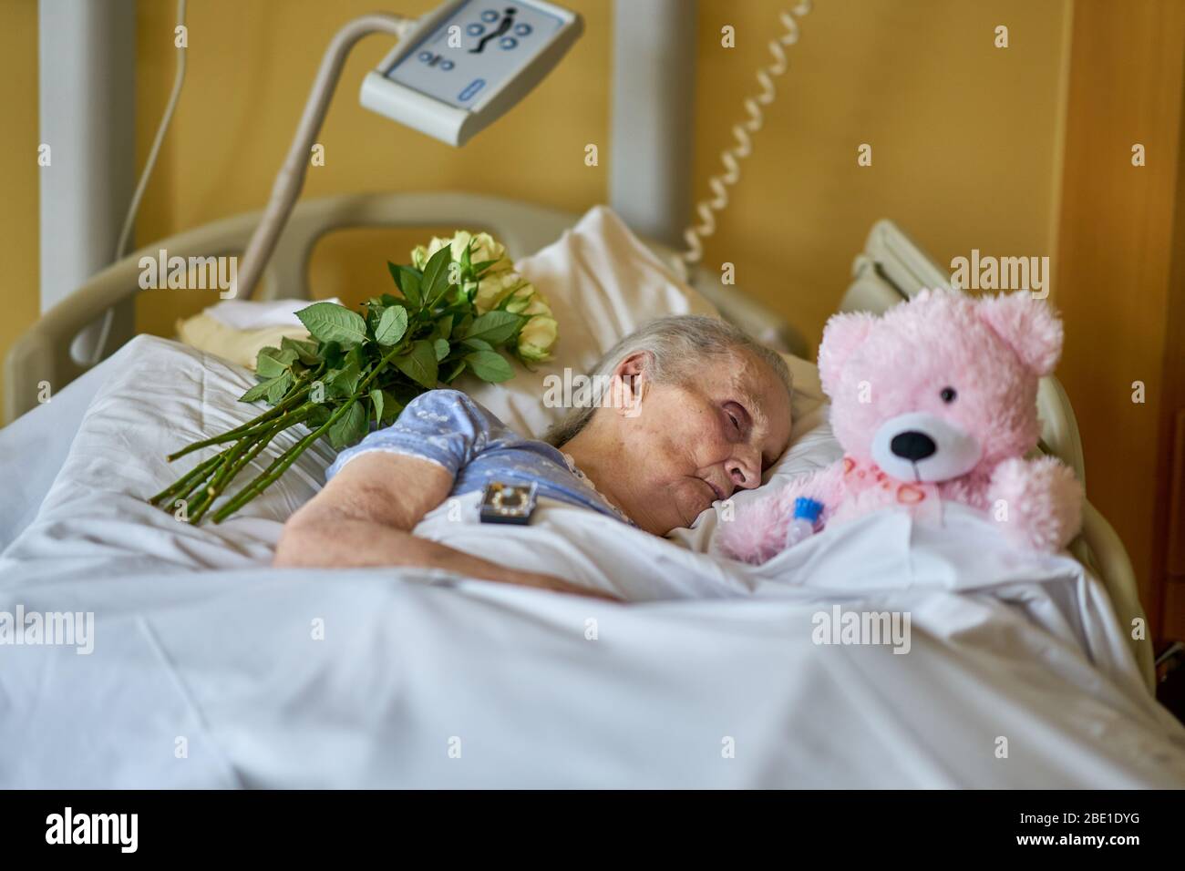 Eine Frau liegt sterbend in einem Krankenhausbett, umgeben von einem einäugigen Teddybär und gelben Rosen. Stockfoto
