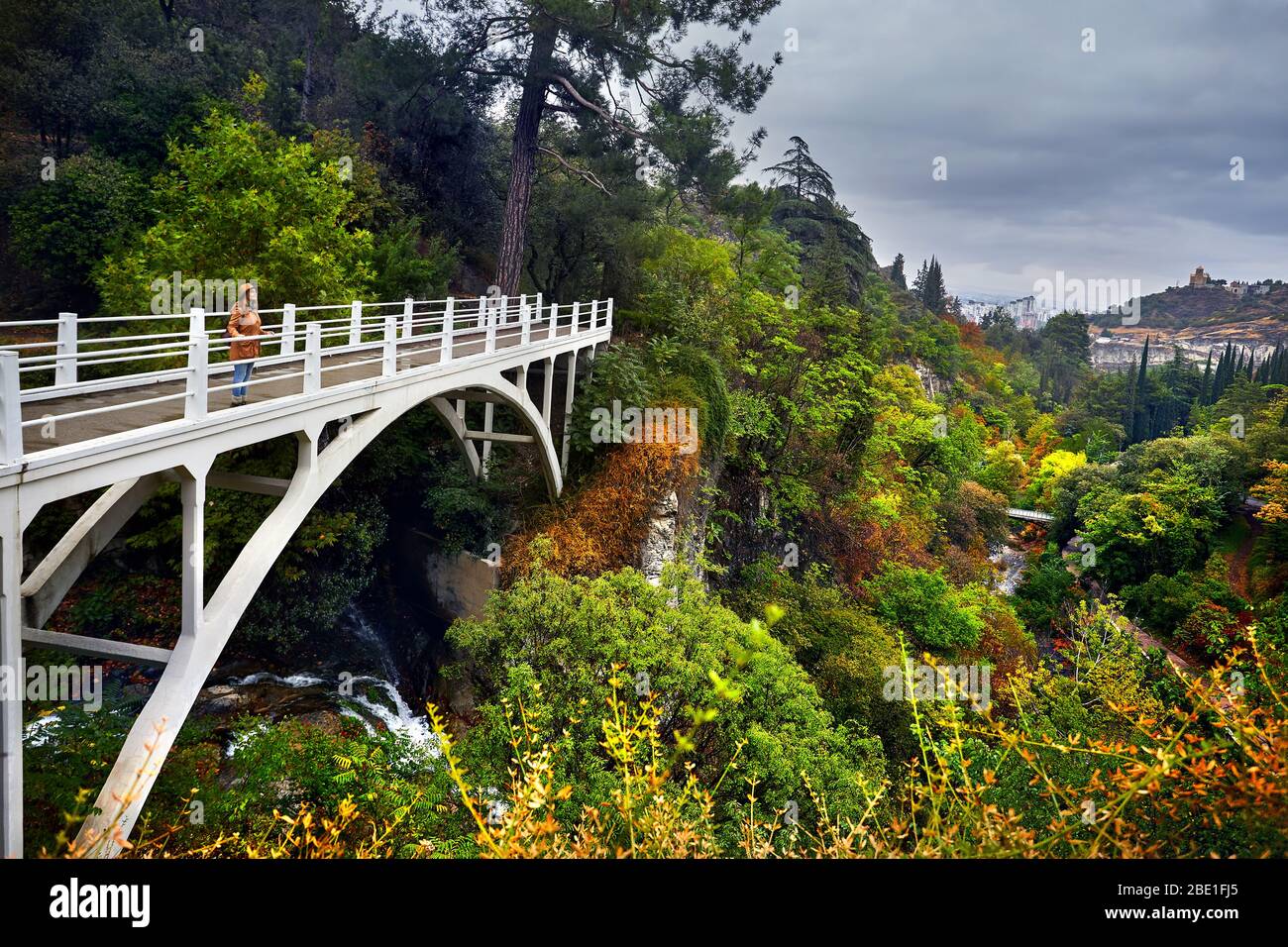 Touristische Frau in Hat an der weißen Brücke im Botanischen Garten mit Bäume im Herbst an bedeckt bewölkten Himmel in Tiflis, Georgien Stockfoto
