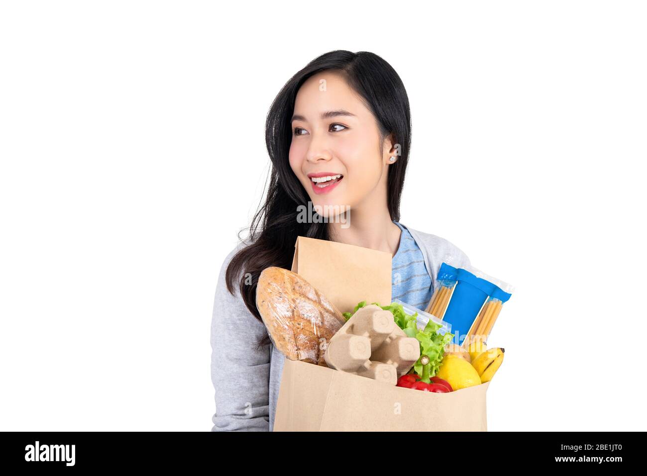 Schöne lächelnde asiatische Frau hält Lebensmitteleinkaufstasche und Blick weg zu leerem Raum beiseite isoliert auf weißem Hintergrund Stockfoto