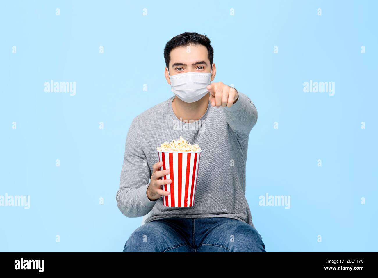 Junger Mann mit Popcorn schützen sich durch das Tragen medizinischer Gesichtsmaske während genießen Film isoliert auf hellblauem Hintergrund Stockfoto