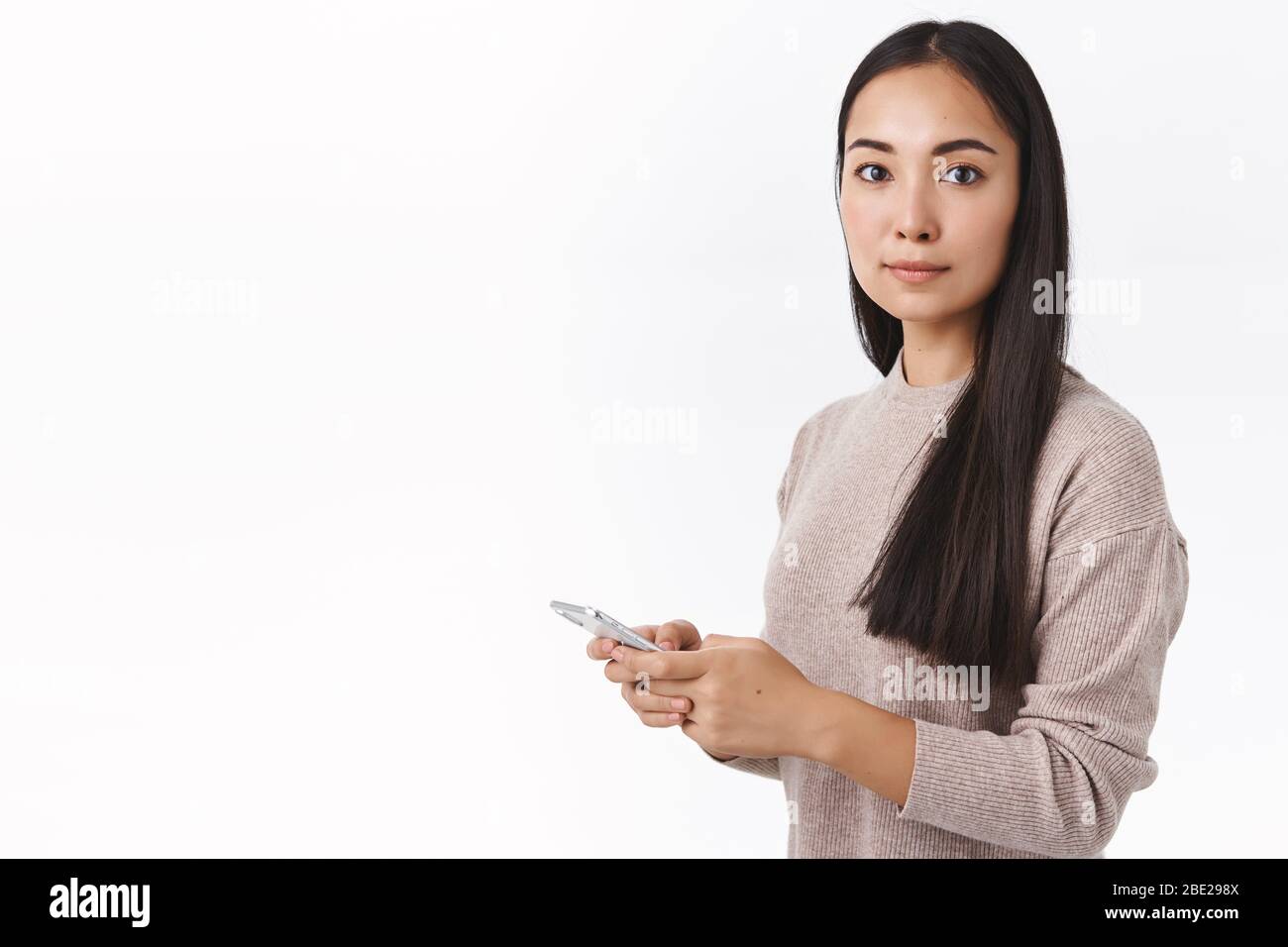 Gut aussehende elegante junge moderne asiatische Frau Mitarbeiterin in beige Pullover, Smartphone halten Kamera drehen fasziniert, hören jemand ihren Namen nennen Stockfoto