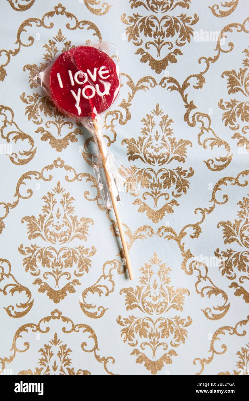Ein süßes gewickeltes 'I Love You' rotes Lollypop vor einem Gold und hellblau dekorierten Hintergrund, flaches Lay, Draufsicht, minimalistisch Stockfoto