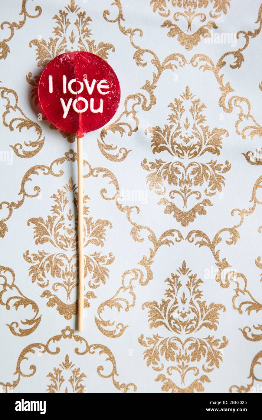 Ein süßes 'I Love You' rotes Lollypop vor einem Gold und hellblau dekorierten Hintergrund, flaches Lay, Draufsicht, minimalistisch, vertikal Stockfoto