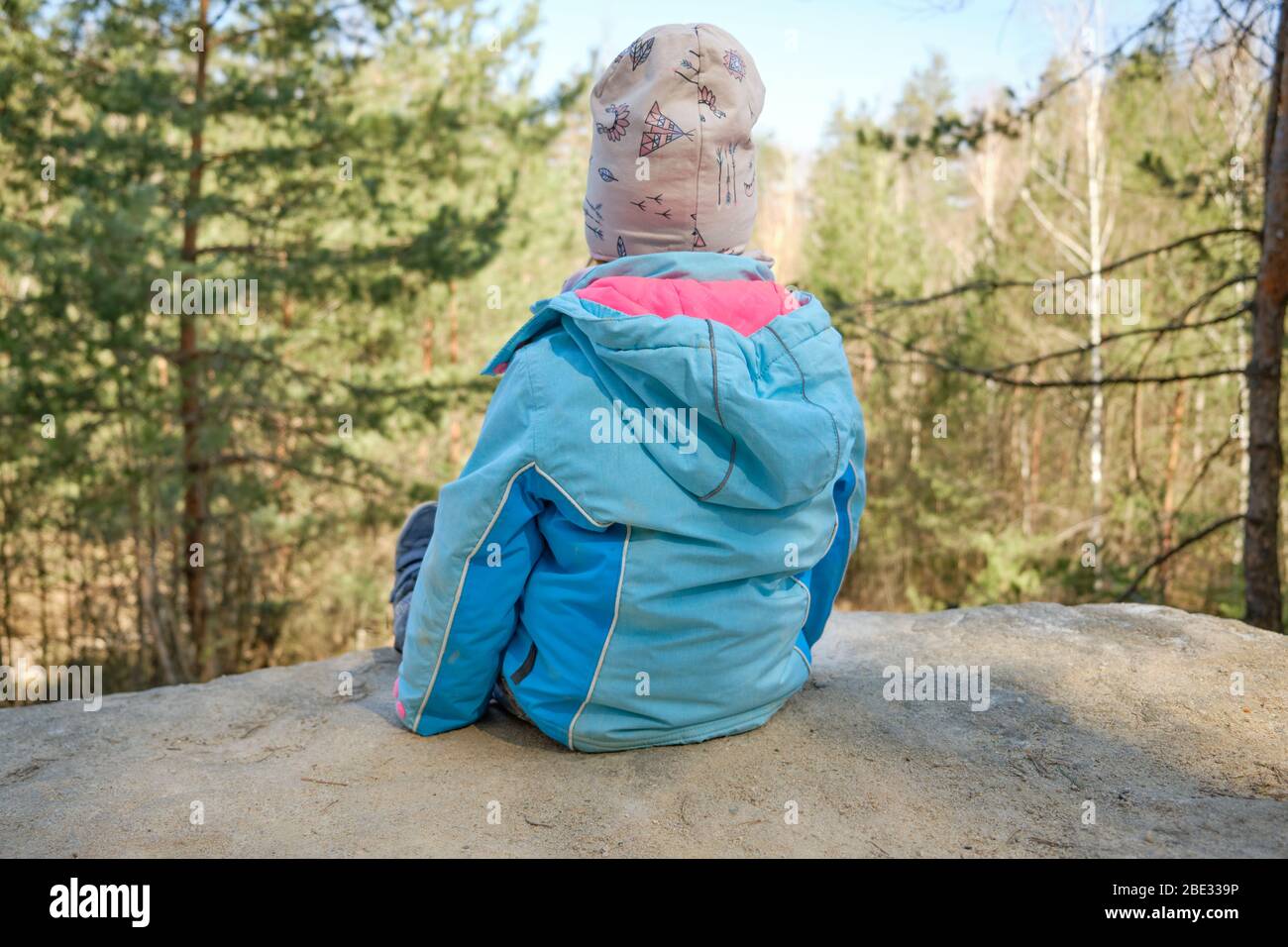 Rückansicht des 4-jährigen kaukasischen Kindes, das auf Sandsteinfelsen sitzt und an einem schönen Frühlingstag in Deutschland den Blick auf den Wald genießt Stockfoto