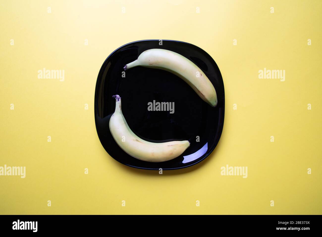 Ein modernes pastellfarbenes Bild von zwei Bananen und ein dekorativer schwarzer quadratischer Teller mit pastellgelbem Hintergrund, flacher Lage, Draufsicht Stockfoto